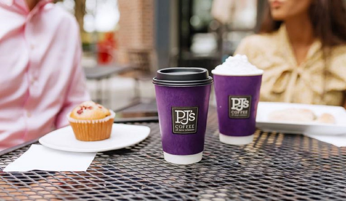 Two PJ's Coffee cups sit on an iron outdoor table in the foreground, with two people and a plate with a muffin on it in the background.
